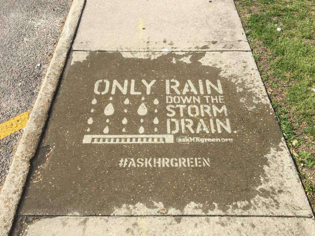 Sidewalk art reads Only rain down the storm drain with images of raindrops and a drain grate, promoting the hashtag #ASKHRGREEN to encourage proper stormwater practices.