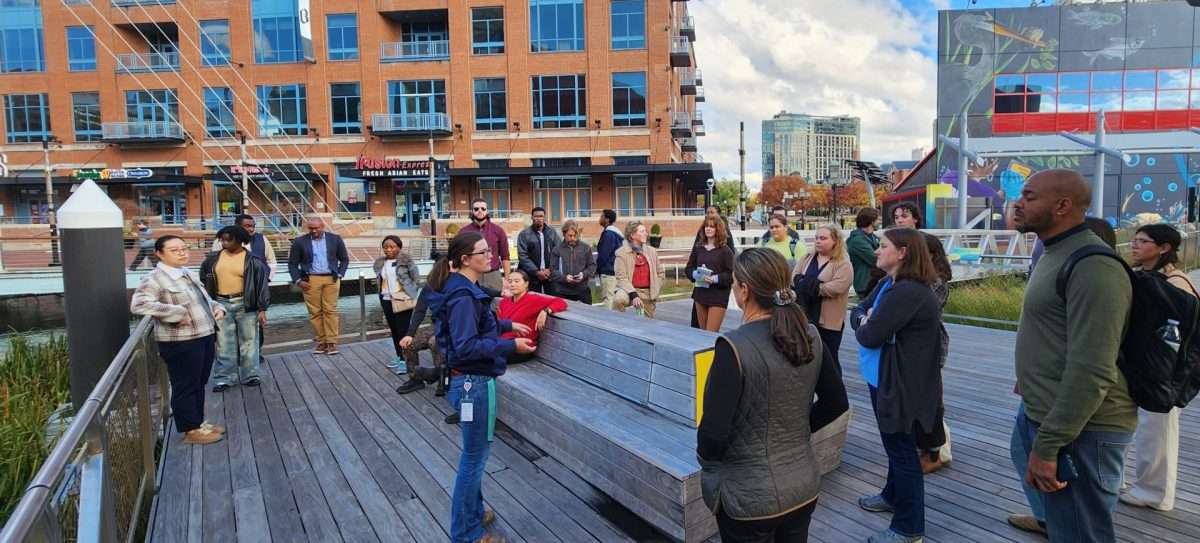 A group of people stands and sits on a wooden boardwalk near the water, listening to a person speaking. Modern buildings and a colorful mural are visible in the background under a partly cloudy sky.