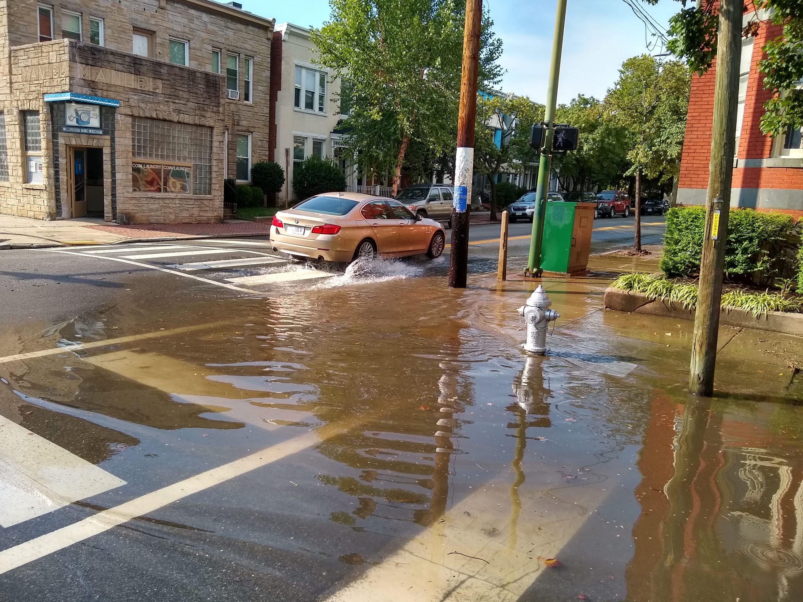 A beige car drives through a flooded intersection, creating splashes, with water pooled around a fire hydrant. Buildings and trees line the street on a sunny day.