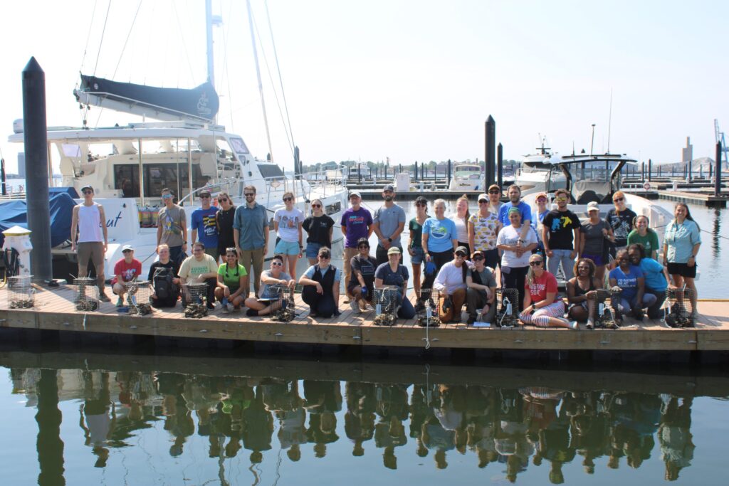 A large group of people pose together on a dock by the water, with boats and yachts in the background on a sunny day. Some people are kneeling and others are standing, and everyone is facing the camera.